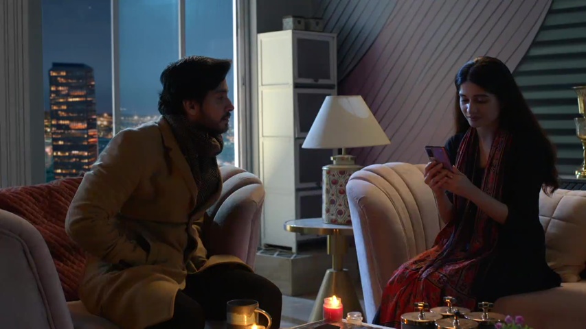 Savi smiling at her phone while Neil lights a candle in the dark